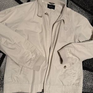 Beige jacka från Oscar Jacobson - Snygg beige jacka från Oscar Jacobson med dragkedja och ribbade detaljer. Jackan har en stilren design med broderad logotyp på bröstet och knappdetaljer vid kragen. Storlek XL tror jag men skulle säga mer som M 