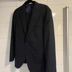 Mörkblå kostym från Pour Homme - Elegant mörkblå (väldigt mörkblå) kostym från Pour Homme i en klassisk design. Tillverkad av en blandning av ull och polyester, vilket ger en bekväm och stilren passform. Perfekt för formella tillfällen. Det hänger byxor innanför kavajen om det inte framgår. STORLEK B96, vilket motsvarar ungefär vanlig kostymstorlek 46, googla vid oklarhet. Jag hade den när jag var storlek S ungefär. Kostymen är använd en eller två gånger och är i superbra skick. 