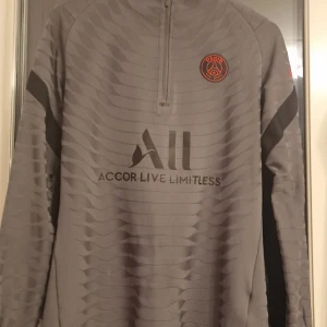 Grå långärmad tröja från Nike - Säljer en grå långärmad tröja från Nike med Paris Saint-Germain-logga på bröstet. Tröjan har en dragkedja vid halsen och texten 'Accor Live Limitless' samt 'Visit Rwanda' tryckt på framsidan och baksidan. Perfekt för fotbollsfans!