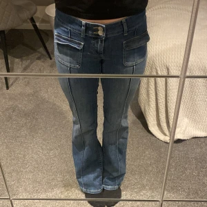 Blå jeans med fickdetaljer - Snygga blå jeans med låg midja och bootcut-stil. De har unika fickdetaljer framtill och en klassisk femficksdesign. Perfekta för en avslappnad look.