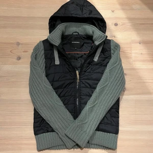 Cardigan - Jacka - Säljer en snygg svart quiltad väst med grå stickade ärmar och huva. Västen har dragkedja framtill och ribbstickade muddar. Perfekt för lager-på-lager under kyliga dagar. Vid minsta fråga eller fundering hör av er🤝