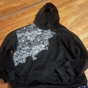 Svart hoodie med mönster - Snygg svart hoodie med ett unikt vitt mönster över ena axeln och bröstet. Tröjan har en klassisk huva och en stor ficka framtill. Perfekt för en avslappnad stil.