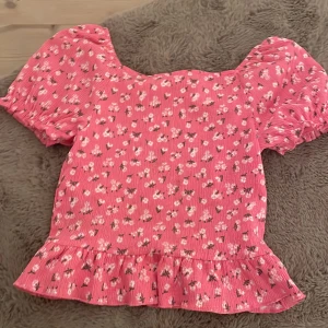 Rosa blommig topp från Kappahl - Söt rosa topp från Kappahl med blommigt mönster och volangdetalj vid nederkanten. Toppen har korta puffärmar och en ribbad struktur som ger en fin passform. 55kr + frakt