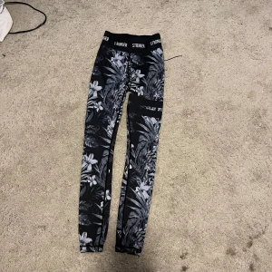 Svarta leggings med blommigt mönster från Stronger - Säljer ett par snygga svarta leggings från Stronger med ett coolt blommigt mönster i grått och vitt. De är högmidjade med en bred resår i midjan där det står 'STRONGER'. Perfekta för träning eller en chill dag hemma. Superbekväma och i bra skick!