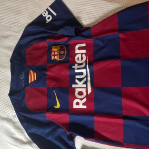 Blå och röd fotbollströja från Nike - Säljer en snygg fotbollströja från Nike i blått och rött med FC Barcelonas emblem på bröstet. Tröjan har korta ärmar och är i Dri-FIT-material för bästa komfort. Den har sponsorer som Rakuten och Beko tryckta på framsidan och är perfekt för alla Barça-fans!