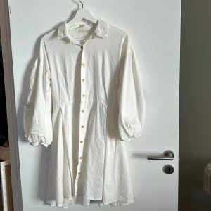 Vit klänning från Foonfie - Säljer en superfin vit klänning från Foonfie. Den har långa ärmar med vackra broderade blommor i färger som svart, orange och rosa. Klänningen har knappar hela vägen ner och en klassisk krage. Perfekt för vår och sommar!