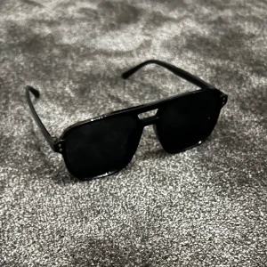 Svarta solglasögon med dubbel brygga - Säljer ett par snygga svarta solglasögon med en cool dubbel brygga. De har en klassisk aviator-stil med mörka linser och en solid svart ram. Perfekta för att ge en stilren look i sommar!