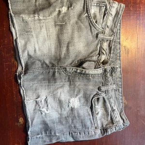 Grå jeansshorts med slitningar - Säljer ett par grå jeansshorts med coola slitningar. De har en klassisk femficksdesign och en knapp- och dragkedjestängning. Perfekta för en avslappnad sommarlook!