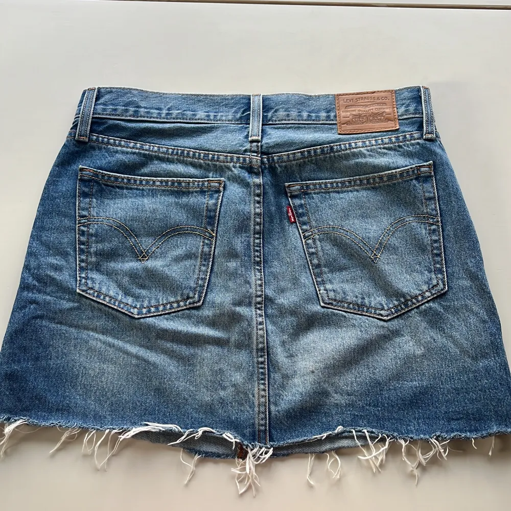 Säljer en snygg jeanskjol från Levi's i storlek M. Kjolen har en klassisk blå denimfärg och är i en kort modell med rå kant nertill för en cool och avslappnad look. Den har fem fickor och en knappgylf. Perfekt för både vardag och fest!. Kjolar.