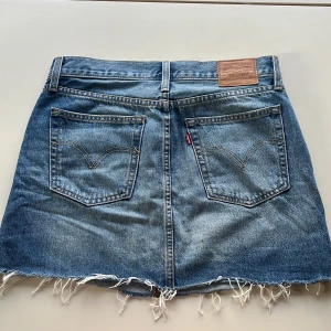 Jeanskjol från Levi's - Säljer en snygg jeanskjol från Levi's i storlek M. Kjolen har en klassisk blå denimfärg och är i en kort modell med rå kant nertill för en cool och avslappnad look. Den har fem fickor och en knappgylf. Perfekt för både vardag och fest!