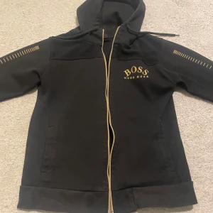 Svart hoodie från Hugo Boss - Säljer en svart hoodie från Hugo Boss med dragkedja och huva. Den har guldfärgade detaljer och logotyp på bröstet. Perfekt för en stilren och bekväm look.