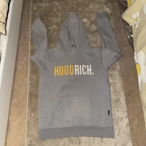 Grå hoodie från Hoodrich - Säljer en grå hoodie från Hoodrich med text i gult och vitt på framsidan. Tröjan har en stor ficka framtill och en bekväm huva. Perfekt för en avslappnad stil. Storlek S men en person som har storlek M kan också ha på sig den.