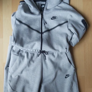 Grå Nike Tech tracksuit - Säljer en grå hoodie från Nike med dragkedja och svarta detaljer. Tröjan har en justerbar huva och är långärmad. Perfekt för en sportig look eller avslappnad stil samt väldigt sköna och stiliga Nik tech byxor!
