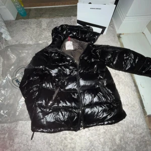 Moncler jacka - Säljer en snygg svart pufferjacka från Moncler med glansig yta och dragkedja. Jackan har en huva och ett unikt märke på insidan. Perfekt för kyliga dagar!
