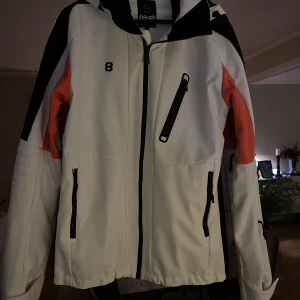 Vit jacka från 8848 Altitude - Snygg vit jacka från 8848 Altitude med svarta och orange detaljer. Jackan har en dragkedja framtill och flera praktiska fickor med dragkedjor. Perfekt för kyligare dagar med sin långa ärmar och huva. I närmaste nyskick då den endast är använd vi en veckas skidresa. 