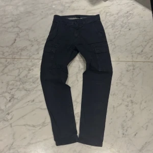 Mörkblå cargobyxor från North Sails - Snygga mörkblå cargobyxor från North Sails med praktiska fickor på sidorna. Byxorna har en dragkedja och knapp framtill för enkel stängning. Perfekta för en avslappnad stil med en touch av funktionalitet. Pris lätt diskuterbart vid snabb affär.