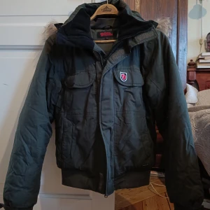 Fjällräven vinterjacka med äkta pälskrage - Mörkgrön vinterjacka från Fjällräven, storlek XS men passar även S. Har en avtagbar krage i äkta päls och flera fickor. Perfekt för kalla vinterdagar!