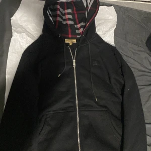 Svart hoodie från Burberry - Säljer en stilren svart hoodie från Burberry med dragkedja och klassiskt rutigt mönster i huvan. Perfekt för en casual look med en touch av lyx. Hoodien har långa ärmar och en bekväm passform. Nyskick inte använt en ända gång pågrund av fel storlek det är XL fast inte i EU storlek den passar mer till M storlek också S Storlek