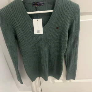 Grön ribbad tröja från Tommy Hilfiger - Snygg grön ribbad tröja från Tommy Hilfiger med v-ringning och långärmad design. Perfekt för en stilren look. Den har en liten logotyp på bröstet som ger en extra touch av stil.