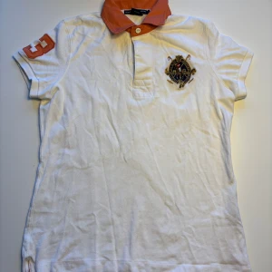 Vit pikétröja från Ralph Lauren - Snygg vit pikétröja från Ralph Lauren, med orange krage och broderat på bröstet. Tröjan har korta ärmar och en klassisk Perfekt för en stilren look!