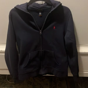 Mörkblå hoodie från Ralph Lauren - Säljer en snygg mörkblå hoodie från Ralph Lauren med dragkedja och det klassiska röda logotypen på bröstet. Tröjan har en bekväm passform och är perfekt för kyligare dagar. Den har en praktisk huva och fickor framtill. Storlek L barn, passar som XS vuxen