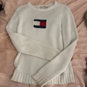 Vit stickad tröja från Tommy Hilfiger - Säljer en stilren vit stickad tröja från Tommy Hilfiger med deras ikoniska logotyp i rött och blått på bröstet. Tröjan har långa ärmar och ribbade kanter vid ärmslut och nederkant. Perfekt för en avslappnad och snygg look.