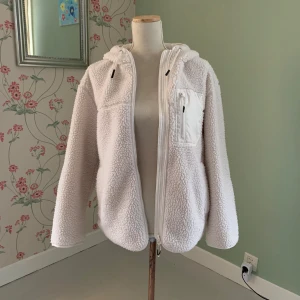 Vit teddyjacka med huva - Mysig vit teddyjacka med huva och dragkedja framtill. Jackan har en praktisk bröstficka med dragkedja och är perfekt för kyligare dagar. Den fluffiga texturen ger en skön och trendig look.