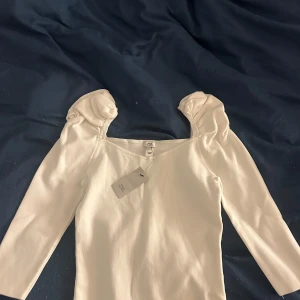 Vit topp från River Island - Säljer en stilren vit topp från River Island med trekvartsärm och puffiga axlar. Perfekt för en chic look. Tillverkad i ett mjukt material som känns skönt mot huden.
