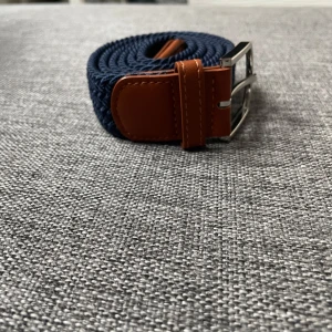 Blått flätat bälte med bruna detaljer - Snyggt blått flätat bälte med bruna läderdetaljer och en stilren silverfärgad spänne. Perfekt för att ge en casual look lite extra stil. Bältet är flexibelt och passar flera storlekar.