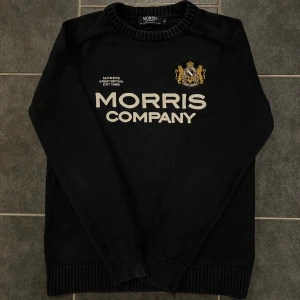 Svart tröja från Morris - Säljer en svart tröja från Morris med texten 'Morris Company' och ett broderat emblem på bröstet. Tröjan är tillverkad i 100% bomull och har långa ärmar samt ribbade muddar vid ärmslut och nederkant.