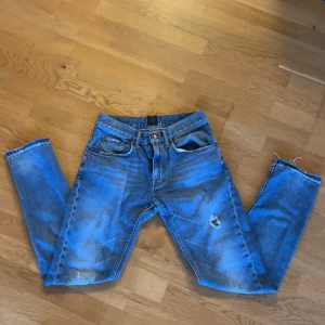 Blå jeans med slitningar - Säljer ett par blå jeans med slitningar och råa kanter. De har en klassisk femficksdesign och en knappgylf. Perfekta för en avslappnad stil med en edgy touch.