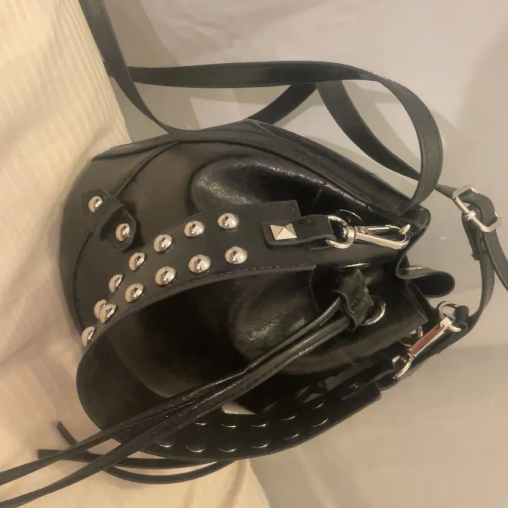Snygg svart axelväska i skinn med coola nitar och justerbar axelrem. Väskan har en unik design med snörning och metallspännen, perfekt för en edgy look. Perfekt för att ge din outfit en extra touch av stil.. Laukut & Käsilaukut.