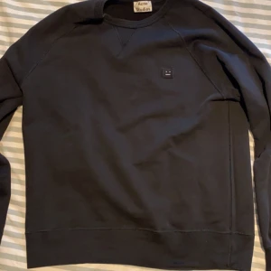 Svart sweatshirt från Acne Studios - Säljer en stilren svart sweatshirt från Acne Studios med deras ikoniska ansiktslogga på bröstet. Tröjan har långa ärmar och en klassisk rund halsringning. Perfekt för en avslappnad och trendig look. Knappt använd inga slitningar eller synliga fläckar.