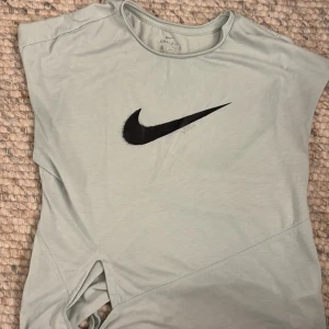 Grön Dri-FIT t-shirt från Nike - Säljer en grön Dri-FIT t-shirt från Nike med svart logga på bröstet. T-shirten har en knytning i sidan och är kortärmad. Perfekt för träning eller en avslappnad stil. 💚