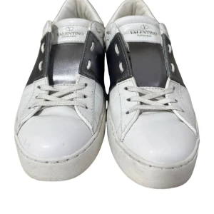 Valentino open - Snygga Valentino Garavani sneakers i vitt skinn med en bred silverfärgad rem över snörningen. Skorna har en klassisk design med rund tå och dekorativa nitar på hälen. Perfekta för en stilren look.