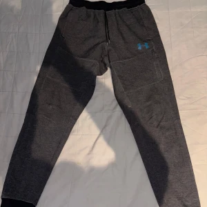 Grå mjukisbyxor från Under Armour - Säljer ett par grå mjukisbyxor från Under Armour med elastisk midja och en liten logga i blått på benet. Byxorna har en avslappnad passform och är perfekta för träning eller en chill dag hemma.