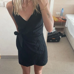 Svart omlottklänning - Snygg svart omlottklänning med knyt i midjan och smala axelband. Perfekt för en stilren look. Klänningen har en kort längd och en v-ringad design som ger en elegant touch.