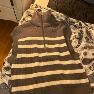 Brun och vit randig ärmlös tröja - Säljer en stilren brun och vit randig ärmlös tröja med dragkedja i halsen. Perfekt för lager-på-lager och ger en avslappnad look. Passar bra till både jeans och kjol.