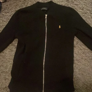  Polo Ralph Lauren Tröja - Säljer en stilren svart tröja från Polo Ralph Lauren med dragkedja och det klassiska broderade logotypen i guld på bröstet. Perfekt för en avslappnad och snygg look. Mycket bra skick och har ett ny pris på ca 2400 men mitt pris 500.