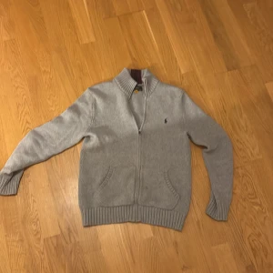 Grå kofta med dragkedja från Ralph Lauren - Snygg grå kofta från Ralph Lauren med dragkedja och ribbade muddar. Perfekt för kyligare dagar. Den har en liten broderad logga på bröstet och en hög krage.