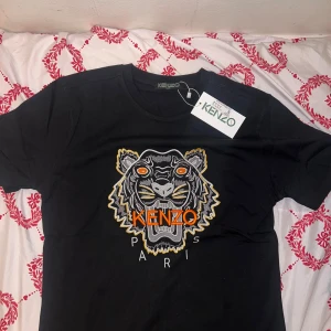 Svart t-shirt med tigerbroderi från Kenzo - Säljer en svart t-shirt från Kenzo med ett coolt tigerbroderi i orange och gult på framsidan. T-shirten har korta ärmar och en rund halsringning. Perfekt för att ge din outfit en edgy touch!