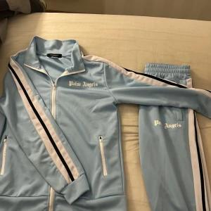 Ljusblå  från Palm Angels tracksuit - Snygga ljusblå  dress från Palm Angels med vita och svarta ränder längs sidorna. Byxorna har elastisk midja och sidofickor. Perfekt för sommaren/våren och inga skador på dem, säljer hela dressen.
