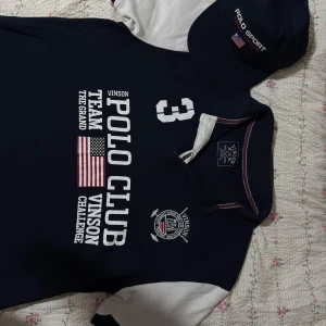 Mörkblå t-shirt från Vinson Polo Club och en keps från Polo sport Ralph lauren - Snygg mörkblå t-shirt från Vinson Polo Club med tryck av amerikansk flagga och text. T-shirten har korta ärmar i kontrasterande vitt och en klassisk krage med röd och vit rand. Perfekt för en sportig look. Kommer också med en polo sport Ralph lauren keps.