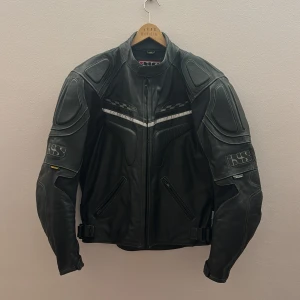 Svart motorcykel jackafrån IXS - Säljer en svart motorcykel jacka från IXS, perfekt för motorcykelentusiaster. Jackan har långa ärmar och dragkedja framtill. Den är designad med skyddande paneler och justerbara remmar i midjan för optimal passform. Märkeslogga syns både fram och bak.