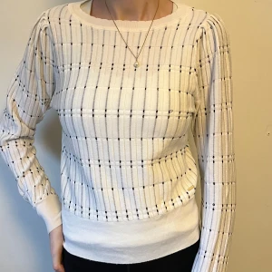 Vit stickad tröja från Lindex - Säljer en vit stickad tröja från Lindex med ett diskret rutmönster i svart. Tröjan har långa ärmar och en rund halsringning. Perfekt för en stilren look.