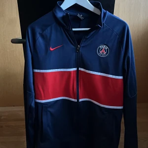 Blå träningsjacka från Nike - Säljer en blå träningsjacka från Nike med röd och vit rand över bröstet. Jackan har dragkedja och långärmade ärmar. Perfekt för sportiga aktiviteter och har en snygg logga på bröstet.