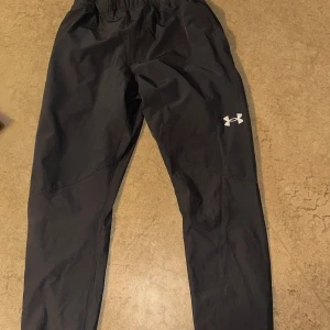 Svarta träningsbyxor från Under Armour - Snygga svarta träningsbyxor från Under Armour med elastisk midja och snörning. Byxorna har en diskret logga på benet och är perfekta för sport och träning. Materialet är lätt och bekvämt för maximal rörelsefrihet.
