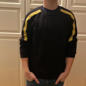 Svart tröja med gula detaljer från EA7 - Säljer en svart tröja från EA7 med gula ränder längs ärmarna. Tröjan har en normal passform och är långärmad, perfekt för en stilren look. EA7-loggan syns på bröstet och längs ränderna.