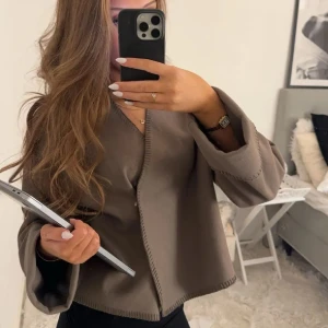 Beige kavaj/kofta - Säljer en stilren beige kavaj/kofta med v-ringad design. Den har långa ärmar och en enkel, elegant look som passar perfekt för både vardag och fest. Kavajen är i bra skick och ger en chic touch till vilken outfit som helst. (Lånad bild)