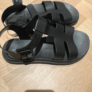 Svarta sandaler från London Rebel - Snygga svarta sandaler från London Rebel i syntetmaterial. De har en stilren design med breda remmar och ett spänne för justerbar passform. Perfekta för sommardagar och ger en cool look till vilken outfit som helst. Använd dem på stranden eller i stan för en avslappnad stil.